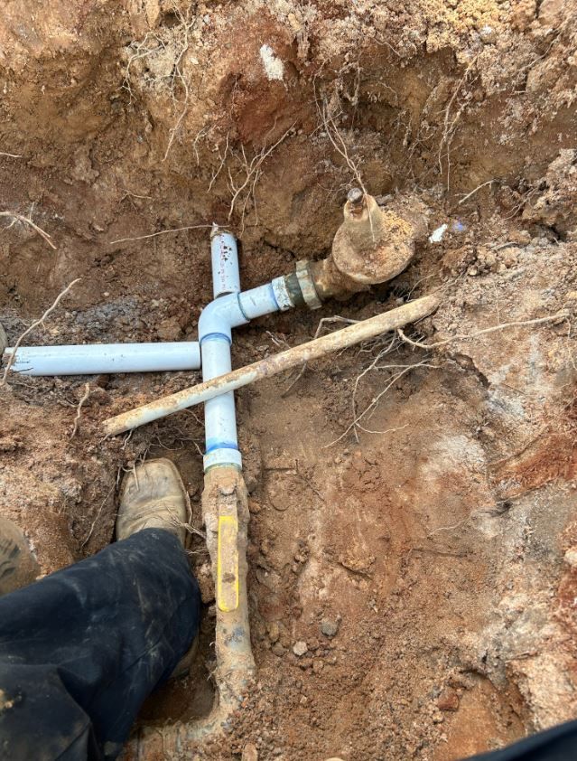 Plumbing pipes and valve exposed in a dirt trench, with a foot visible near the bottom.