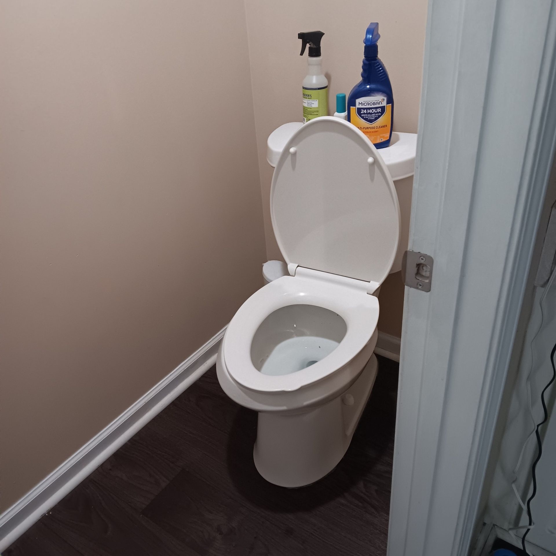 White toilet with open seat, cleaning supplies on the tank, dark flooring, light-colored walls.