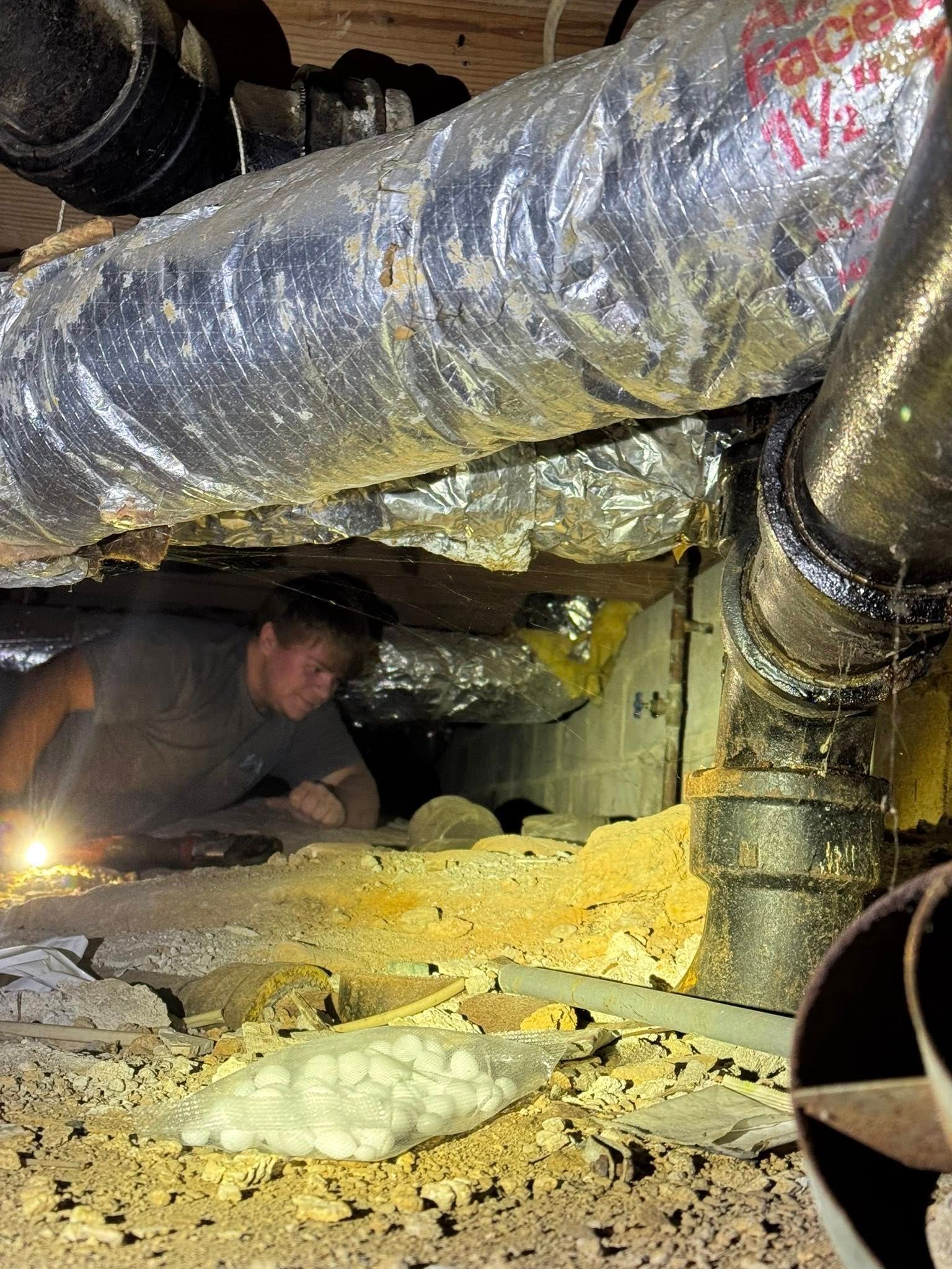 A person crawling in a crawlspace with HVAC ducts and pipes. There is insulation and debris on the floor.