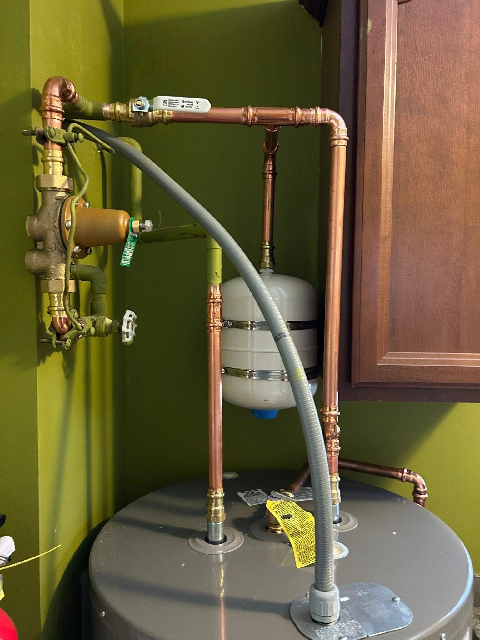 Copper pipes and water heater with expansion tank against a green wall, near a brown cabinet.
