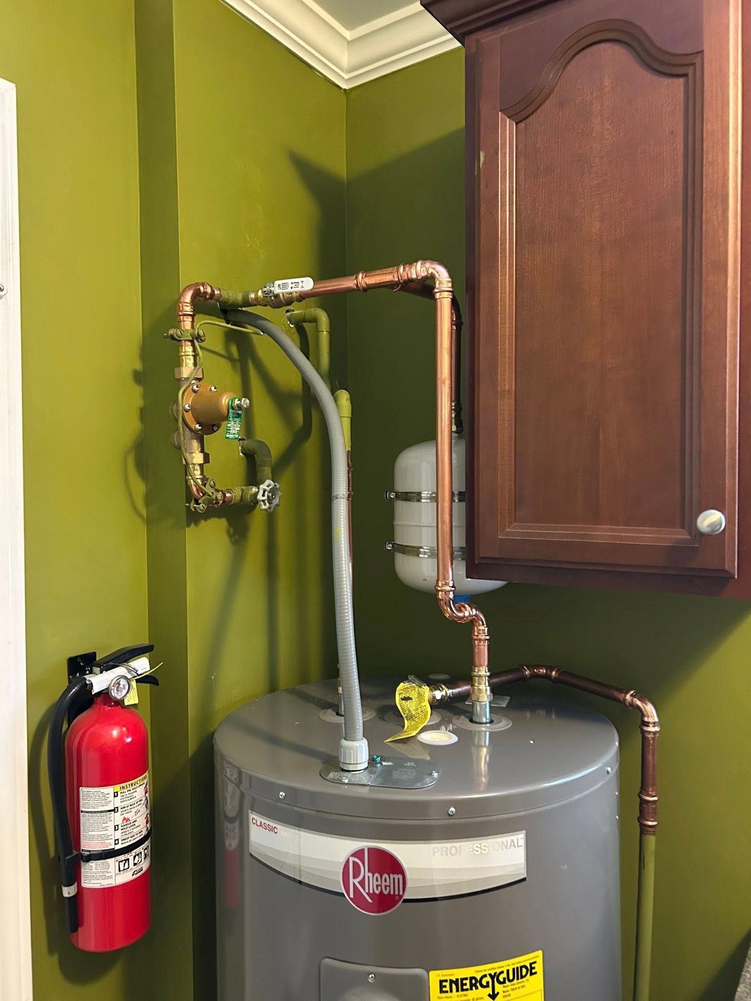 Water heater with copper pipes, tank, pressure relief valve, and a fire extinguisher mounted on a green wall.