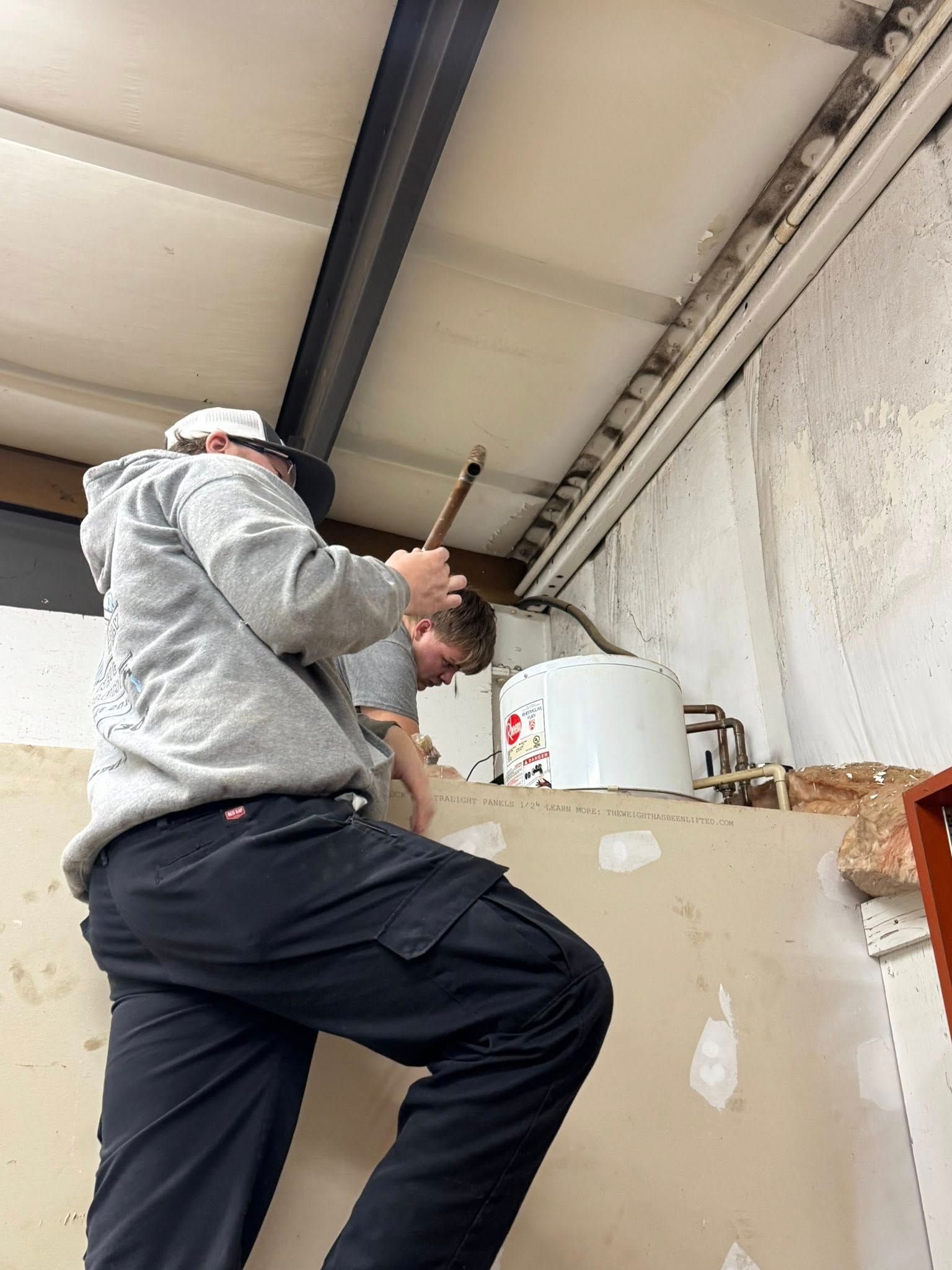 Person in grey hoodie and black pants on a step stool, working on a ceiling, another person assists.