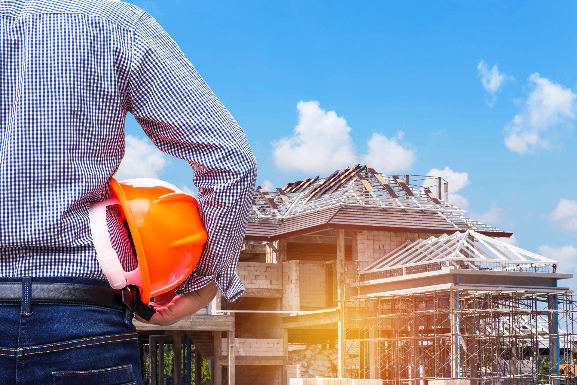 Construction worker holding orange hard hat, overlooking a house under construction on a sunny day.
