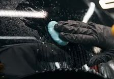 Gloved hand using a blue clay bar to clean a black car's surface. Water spray present, detail shop setting.