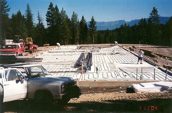 Concrete foundation construction site with workers, trucks, and forested mountains in the background