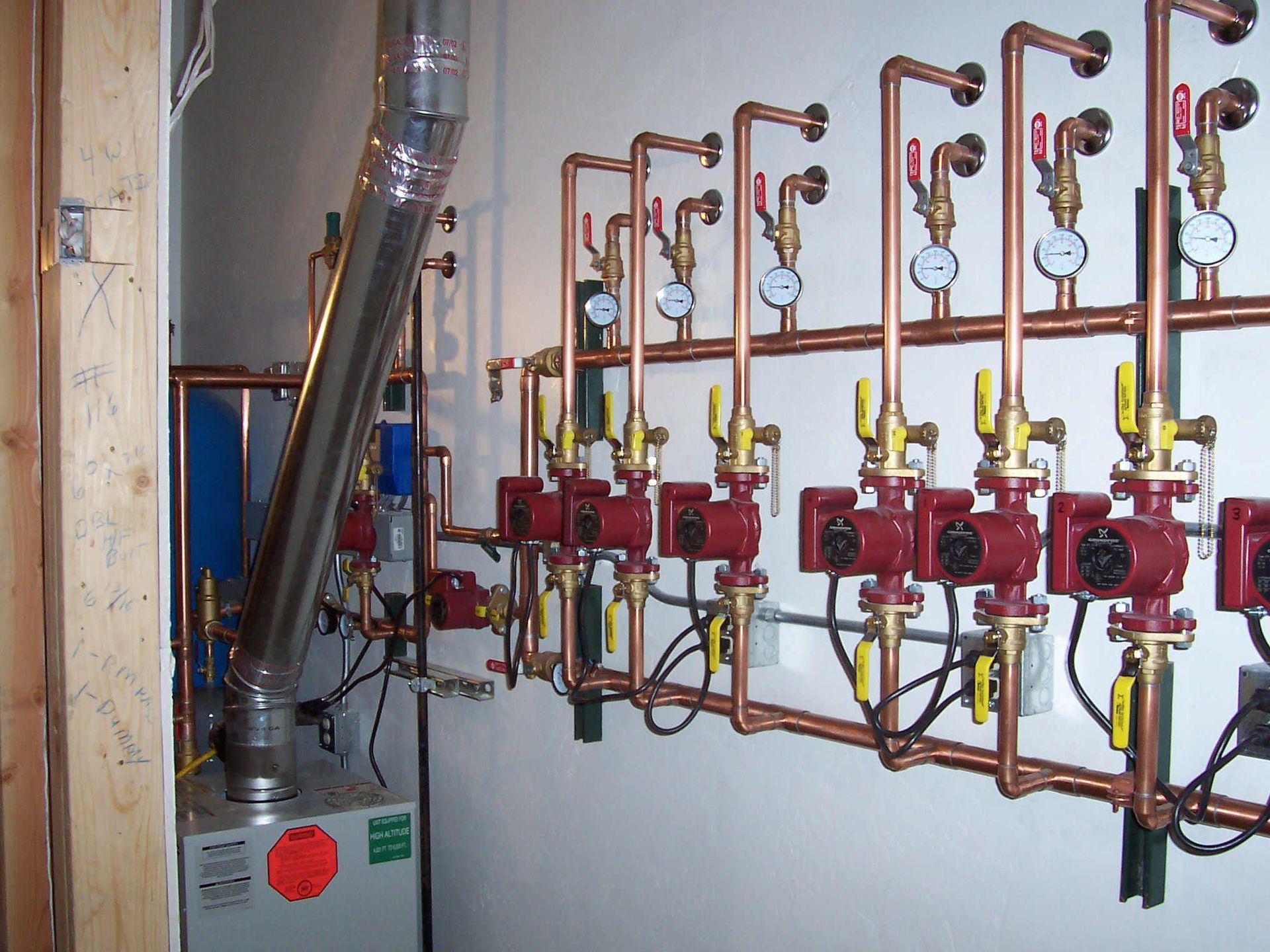 Boiler room with gas pipes, valves, and gauges on a wall, plus a metal flue and heater unit