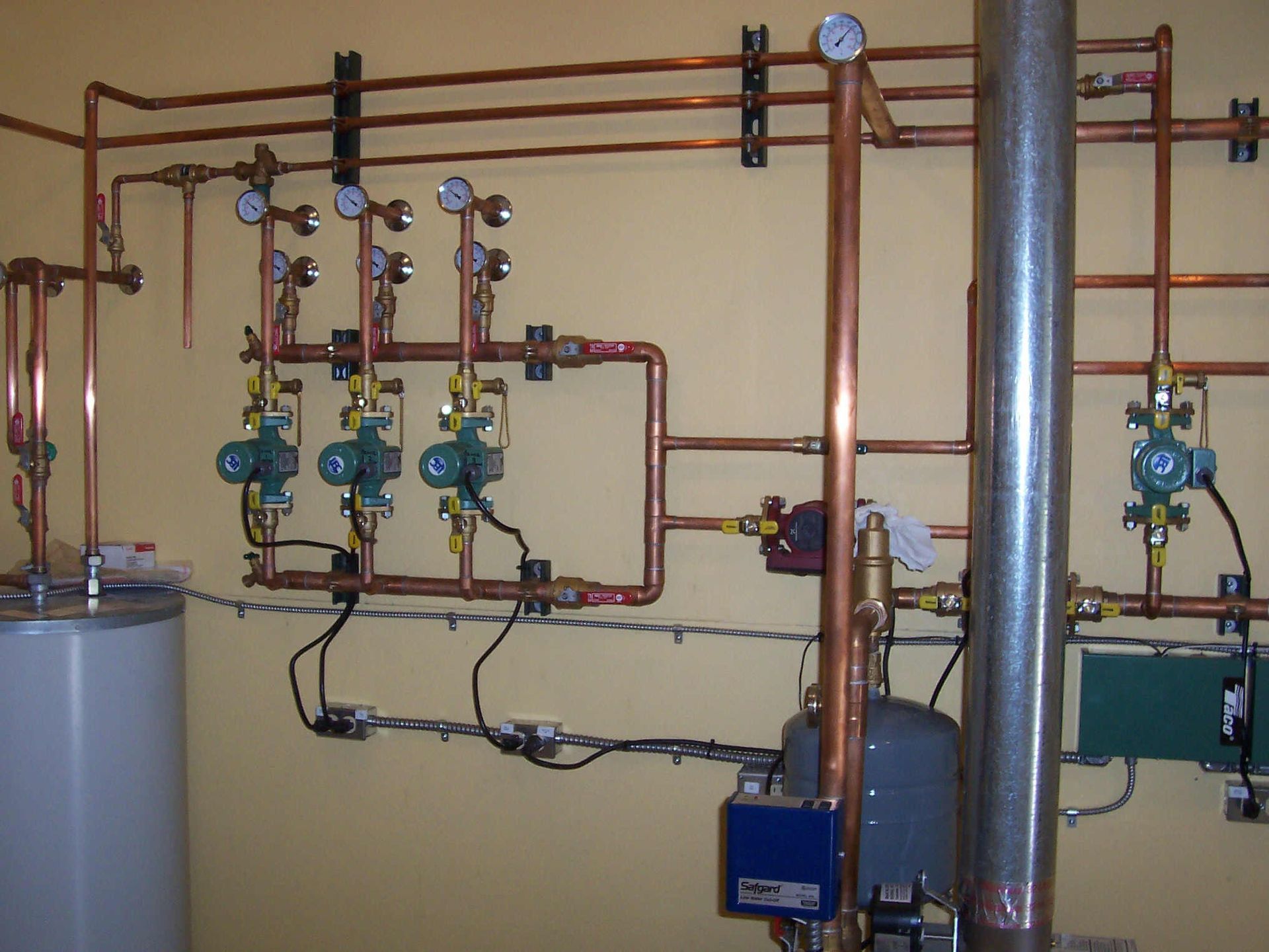 Boiler room with copper pipes, valves, gauges, and a large silver duct beside a blue pump unit