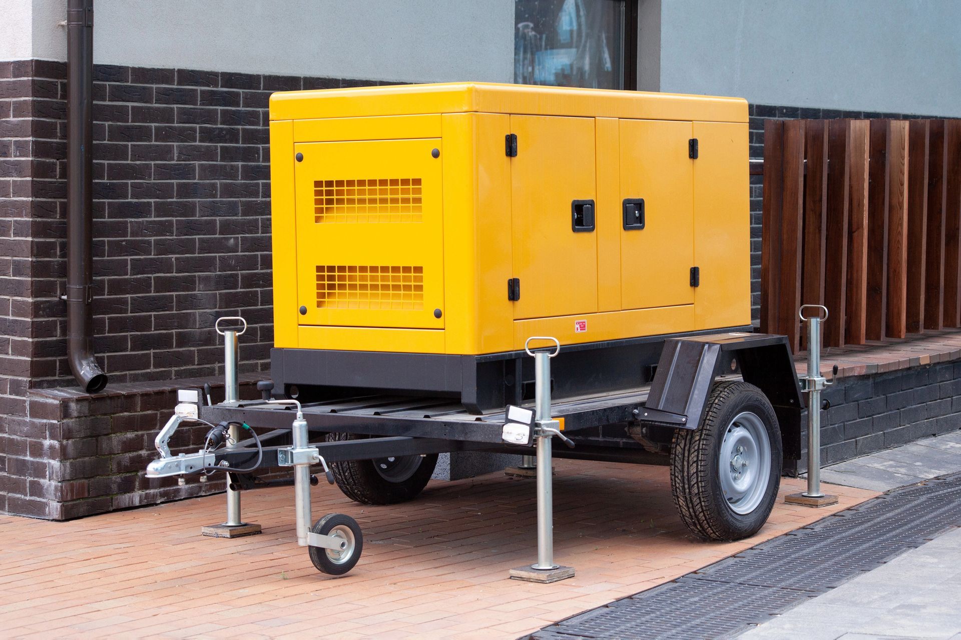Un generatore portatile giallo, montato su un rimorchio nero, è parcheggiato su una superficie pavimentata accanto a un edificio in mattoni.