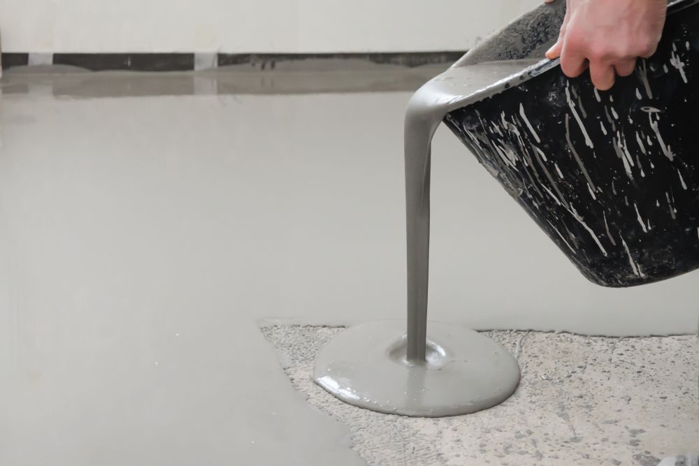 A person pours gray self-leveling compound from a black bucket onto a concrete floor — Western Epoxy Works In Mudgee, NSW