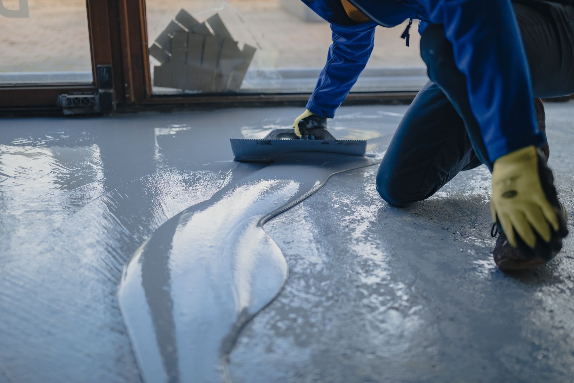 A Worker Kneels on a Floor, Using a Trowel to Spread Gray, Wet Leveling Compound — Western Epoxy Works In Narromine, NSW