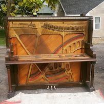 Open upright piano with exposed strings and wooden interior, sitting outdoors on a sidewalk