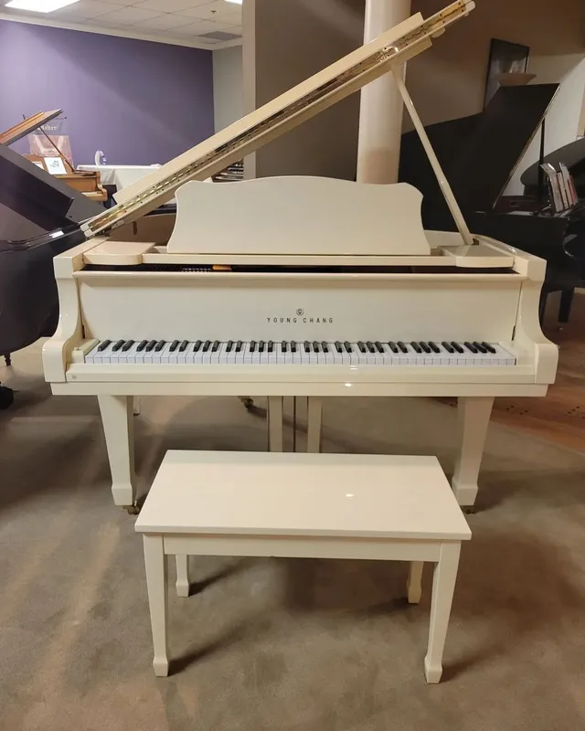 White grand piano with bench in a showroom, lid open, against a purple wall.