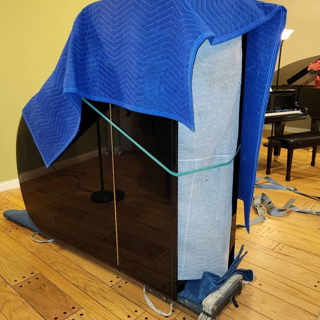 Gray cabinet on a hardwood floor covered with a blue moving blanket, strapped for protection near a piano.