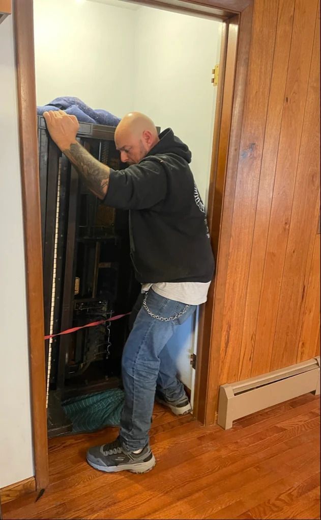 Man working in a narrow closet or utility space, leaning into shelving while wearing a black hoodie and jeans