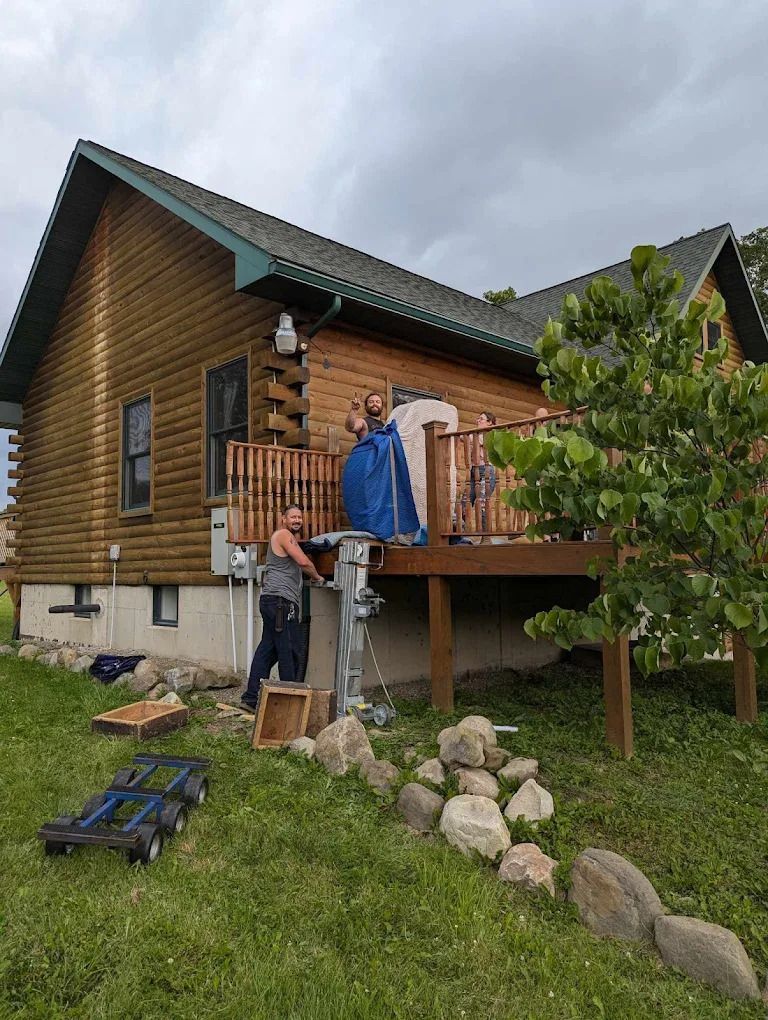 Person standing on wooden deck beside a log cabin, with clothes hanging and rocks in a grassy yard.