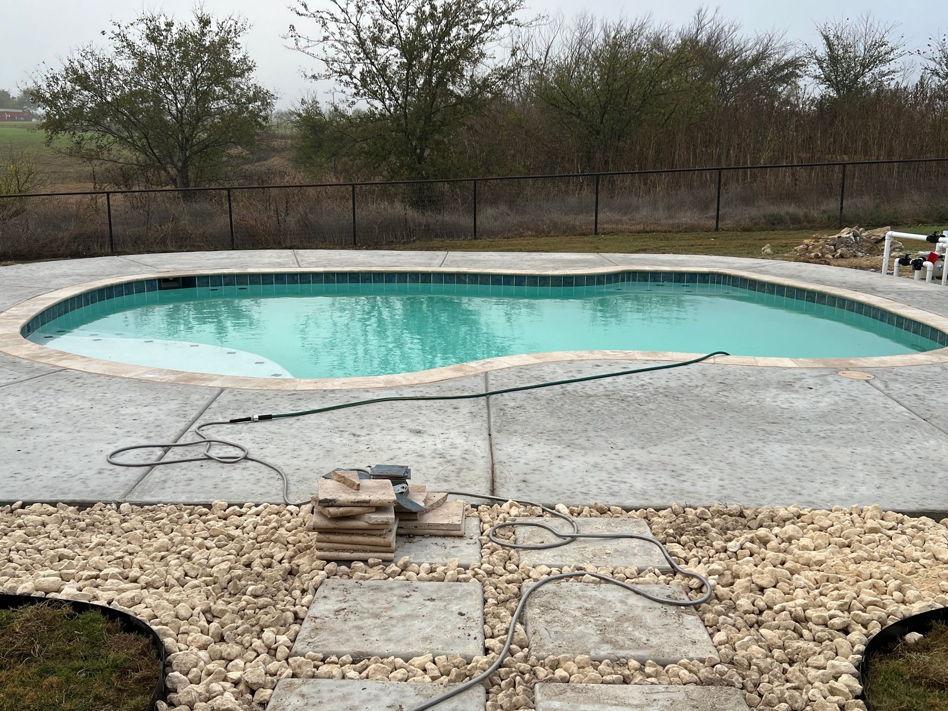 A large swimming pool is surrounded by gravel and a fence.