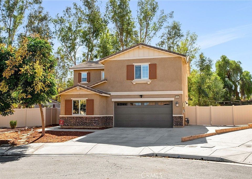 A two-story tan suburban house with a brown garage door, stone accents, and a front yard surrounded by a beige fence.