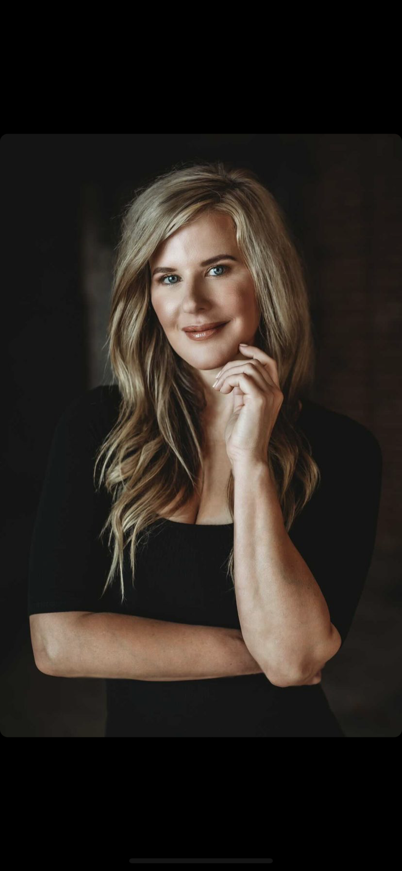 Portrait of a smiling blonde woman in a black top, posing against a dark background