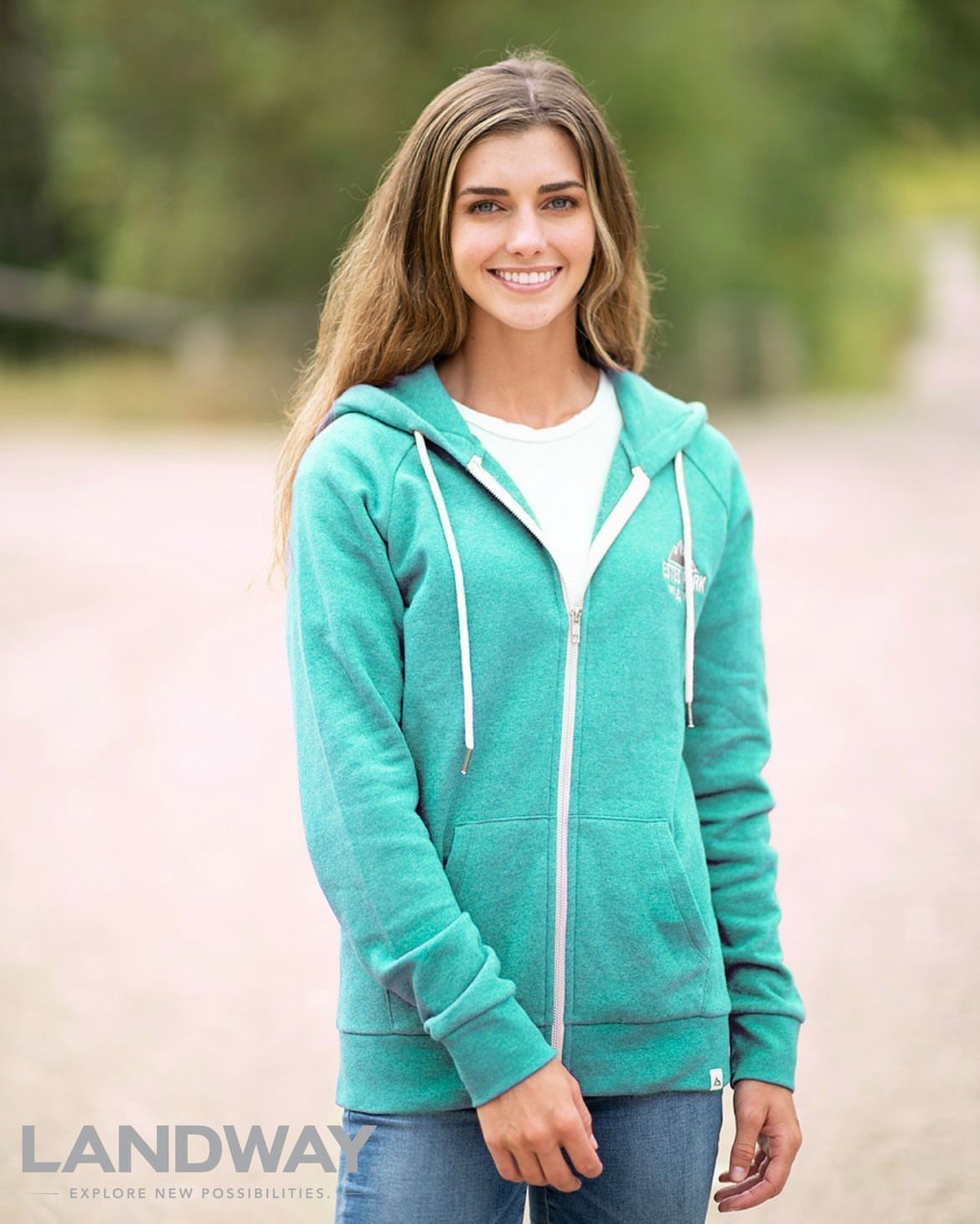 Woman in a teal hoodie and jeans smiling outdoors on a blurred path.