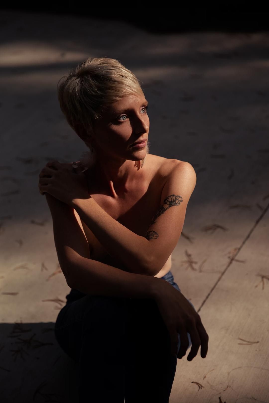 Person with short blond hair seated in dramatic light, arms crossed, against a dark background.