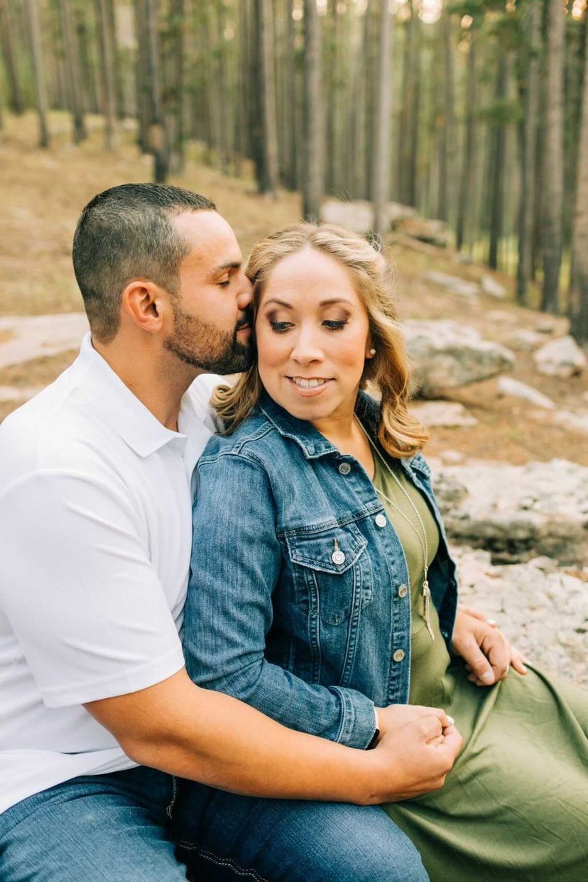 Couple sitting outdoors in a forest, embracing as the man kisses the woman's cheek.