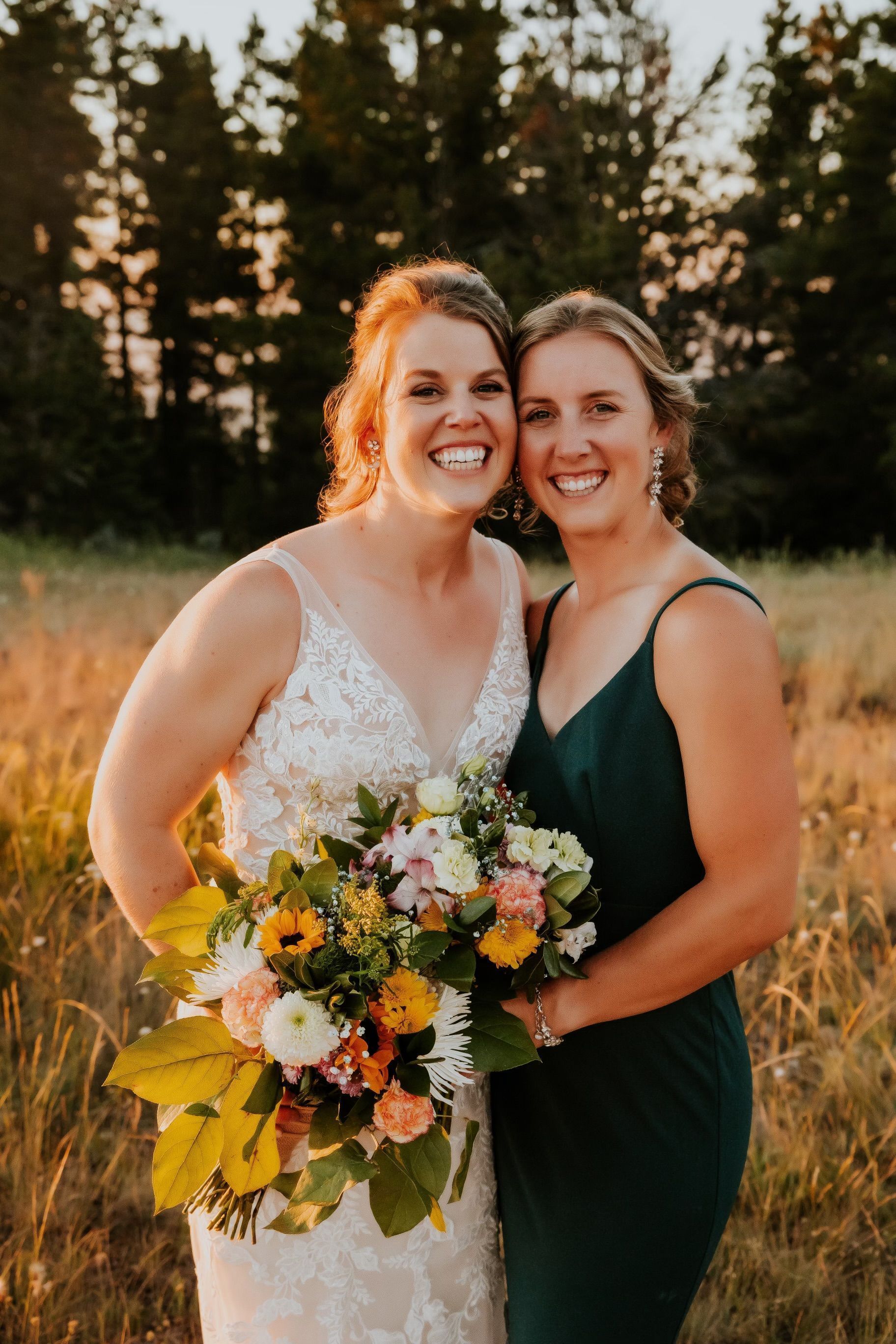 Two women in dresses smiling and holding a bouquet outdoors at sunset