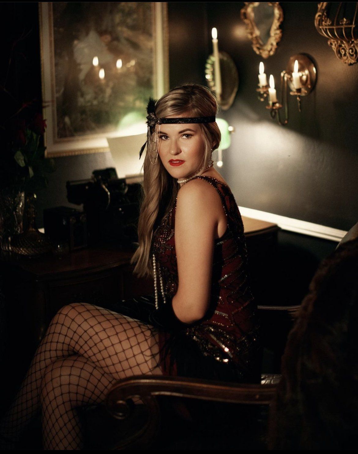 Woman in black fishnet dress and cap seated in a dimly lit lounge, looking over her shoulder