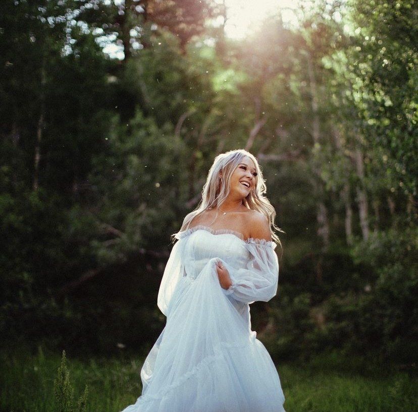 Woman in a flowing white dress standing in a sunlit forest clearing