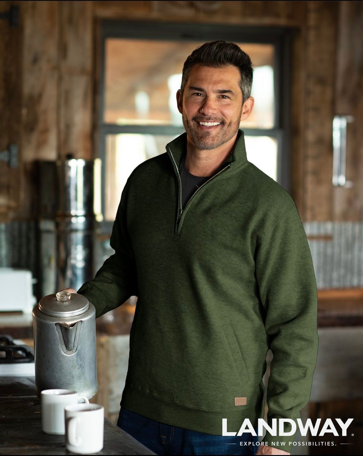 Man in a green sweater smiling indoors beside a metal coffee pot on a counter