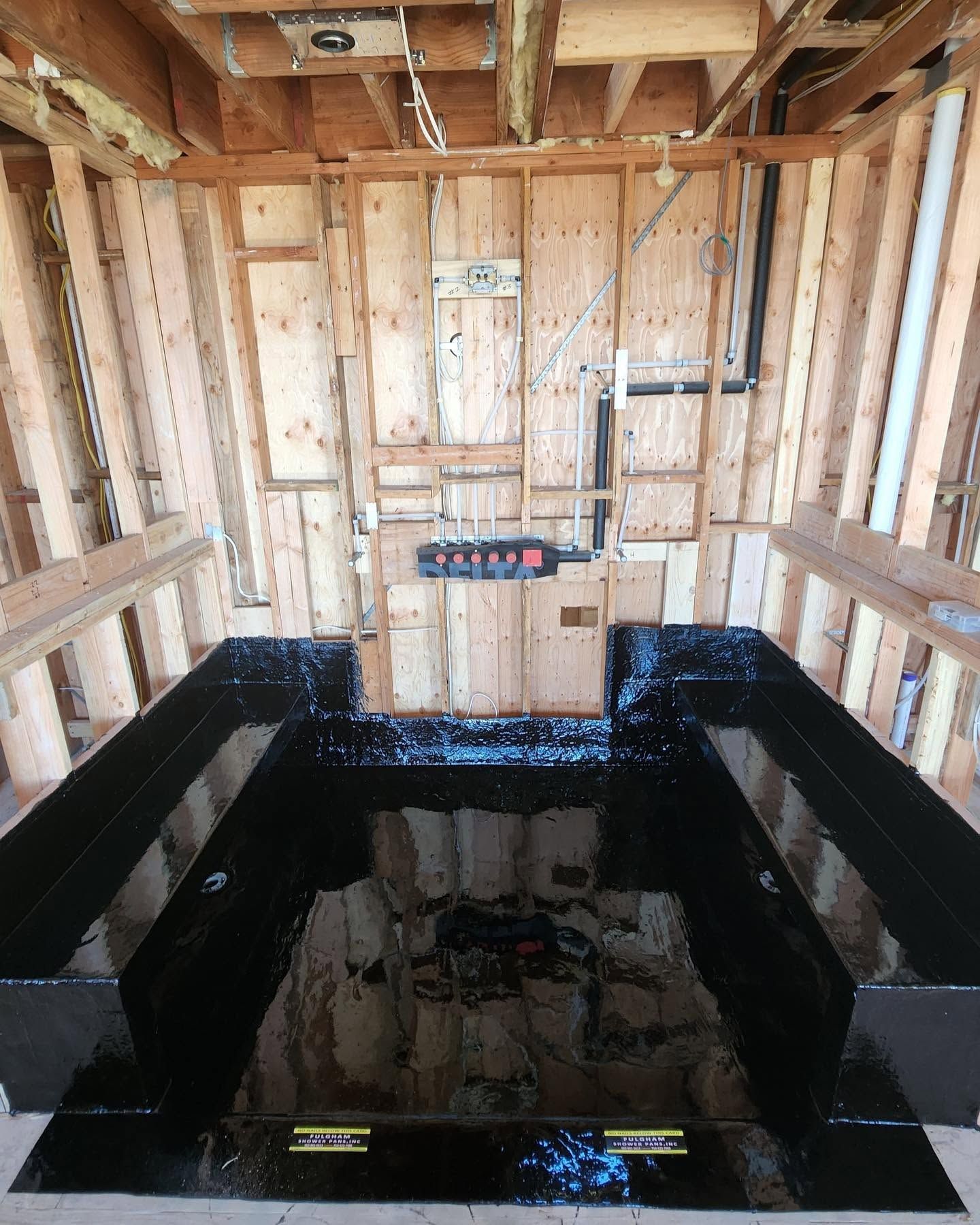 Unfinished wood-framed room with a black waterproofed shower basin and exposed plumbing