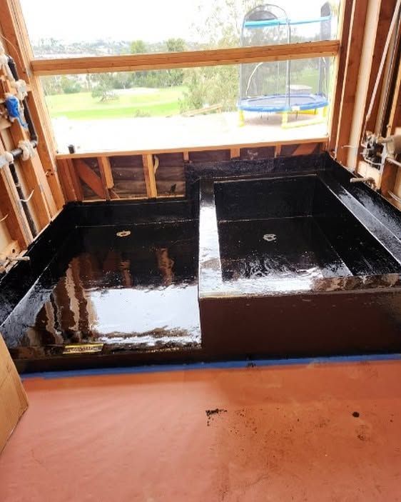 Black waterproofed floor area inside a framed room under construction, with a window overlooking a yard.