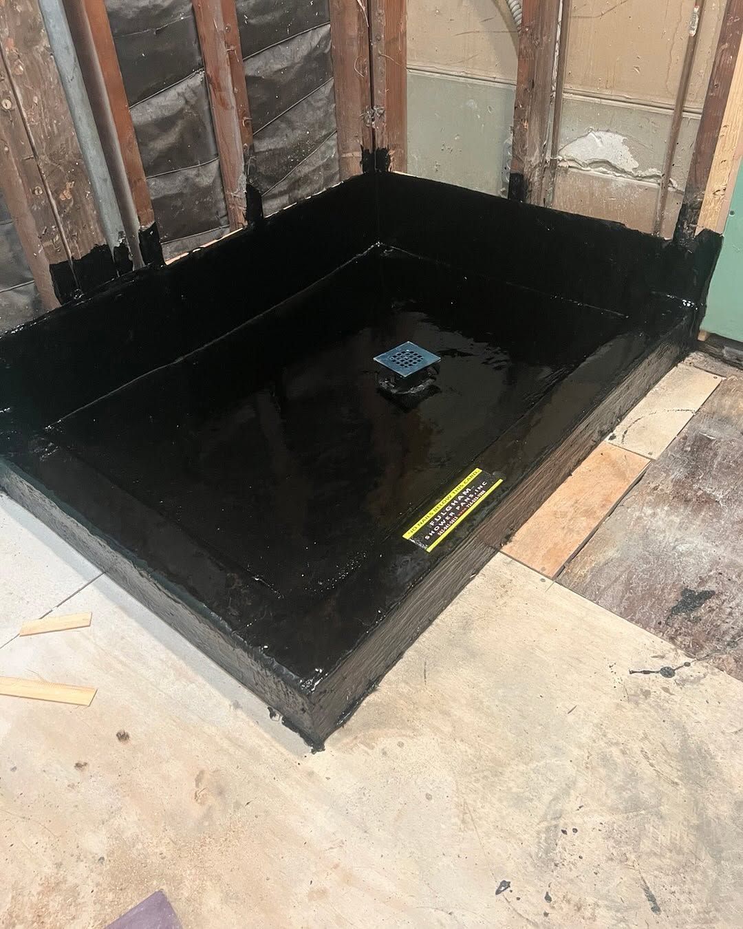Black waterproofed corner pit with a small drain fixture in a partially finished room.