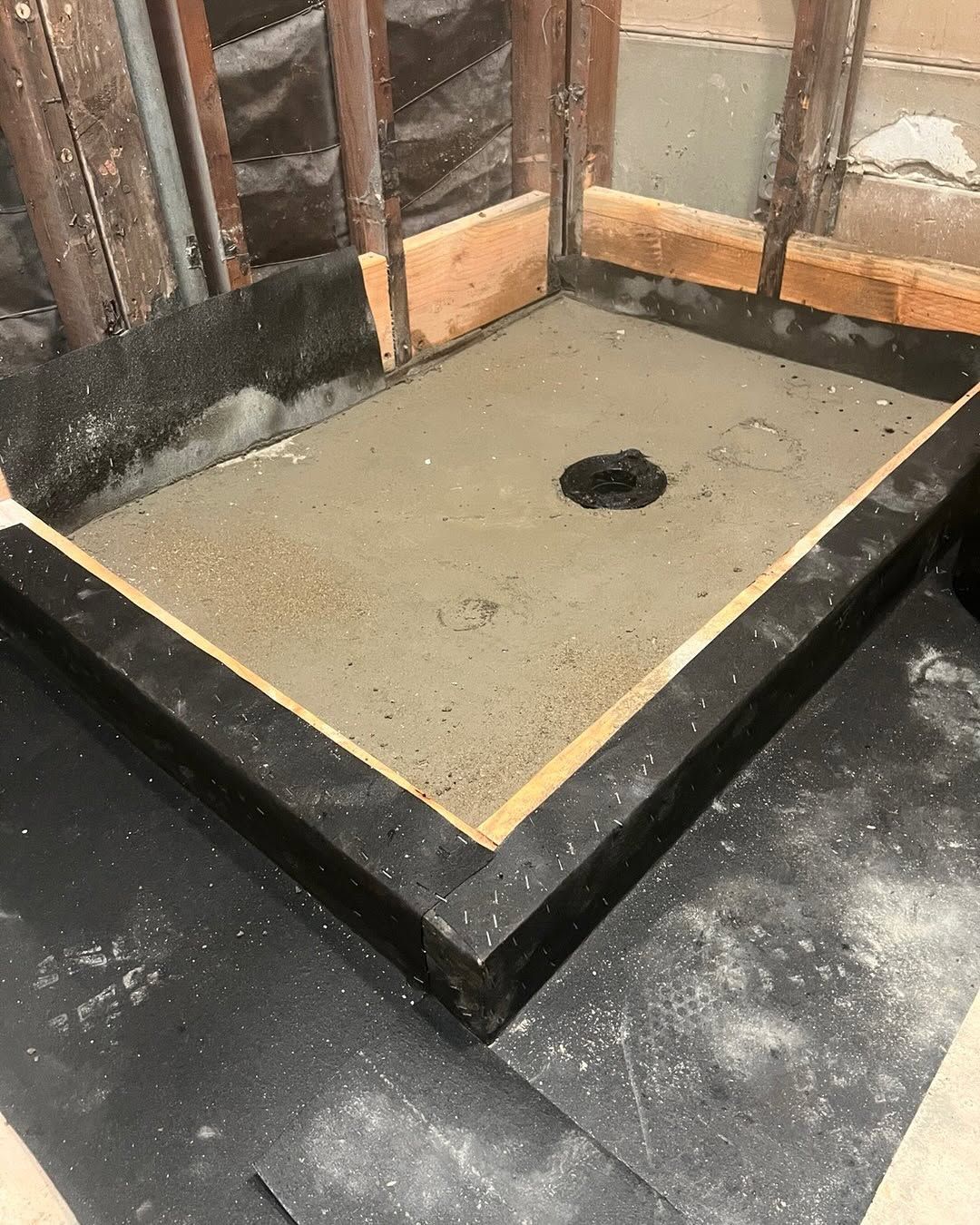 Concrete shower pan under construction with a central drain, surrounded by black waterproofing.