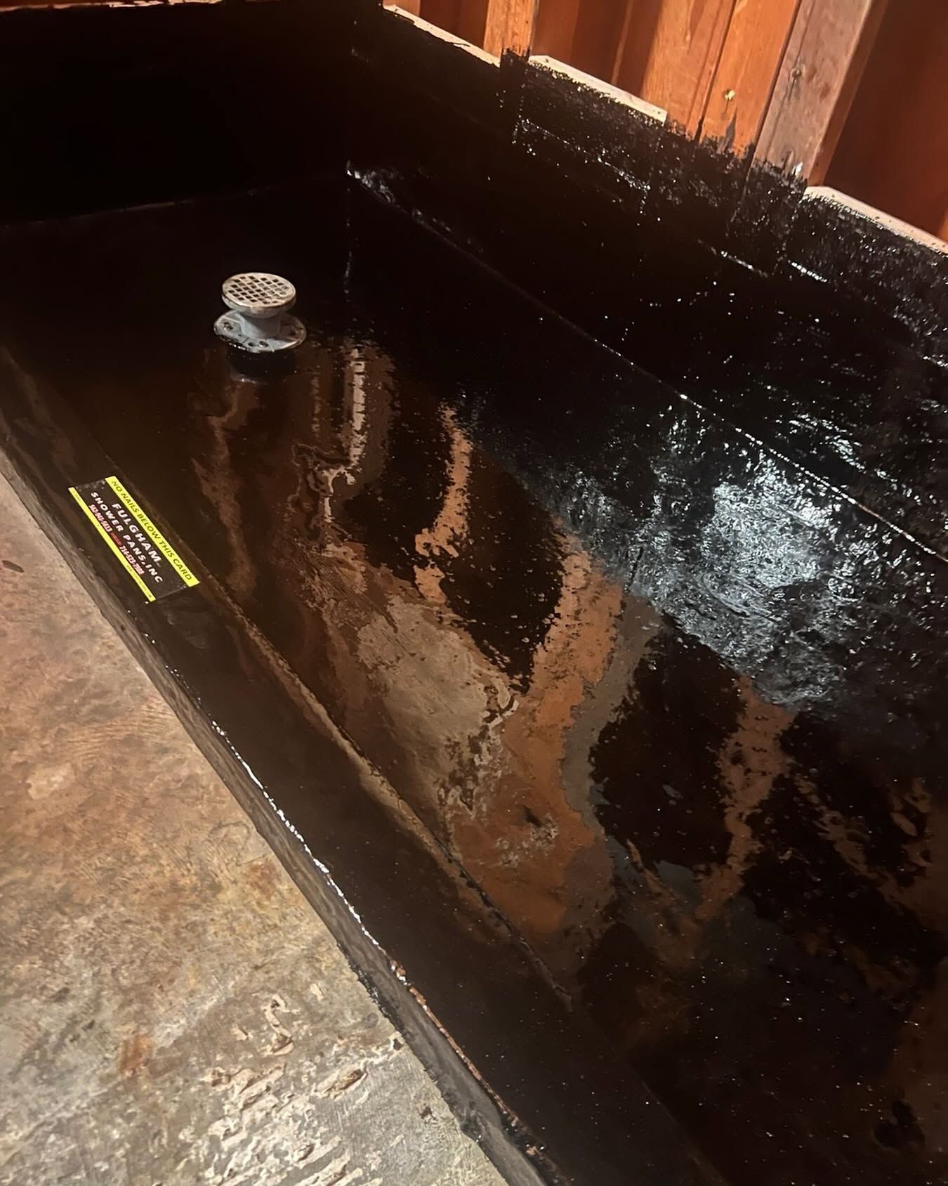 Black waterproof-lined corner in a wooden room, with a small drain or fixture near the wall.