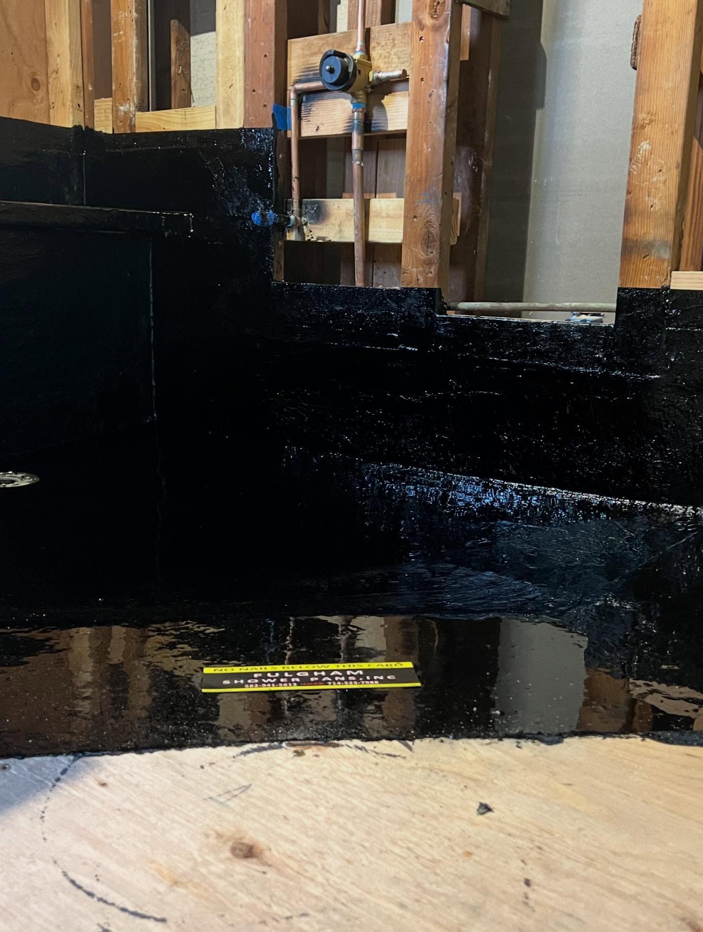 Shiny black waterproofing on wall studs above a bare plywood floor in a construction area