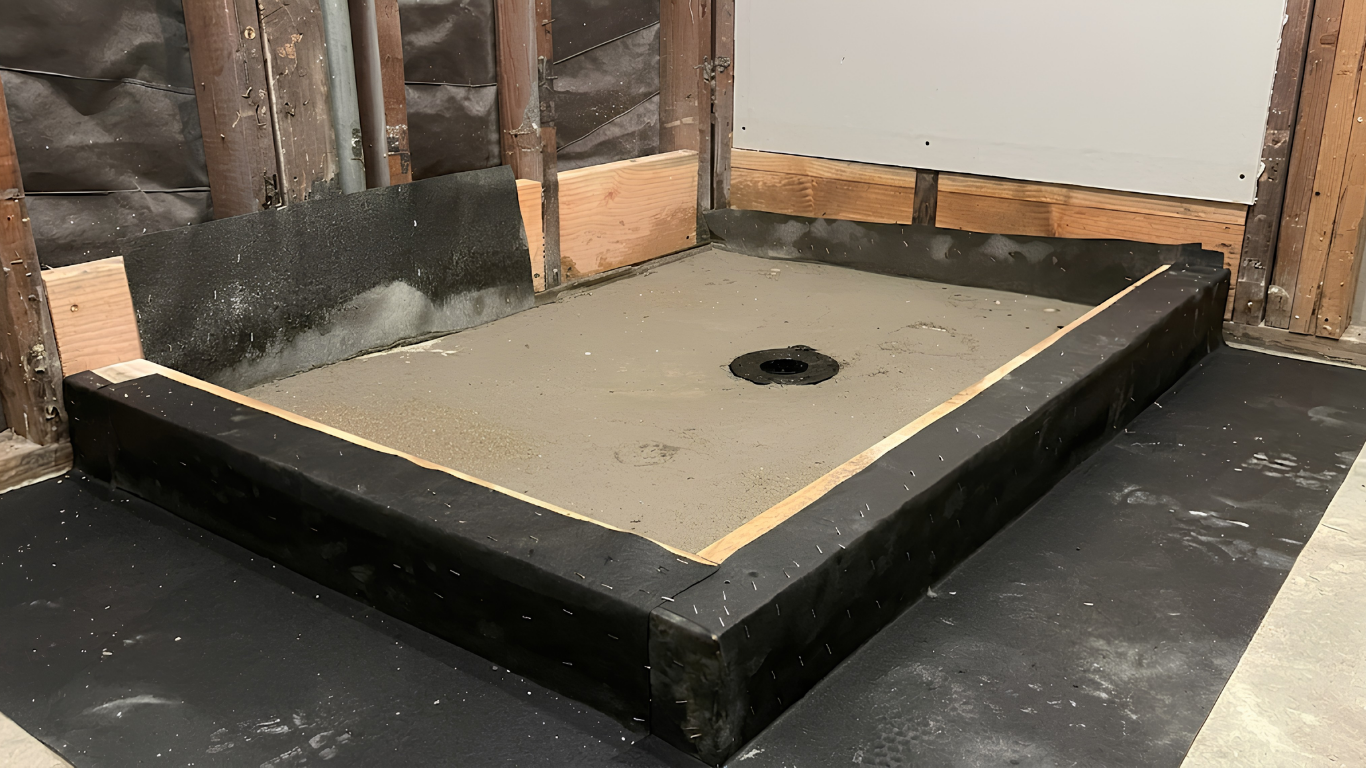 Partially built shower base with black curb, gray floor, and central drain hole in unfinished room
