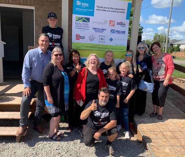 Mikmada Homes team posing in front of a Habitat for Humanity sign. Some give thumbs up. Sunny day.