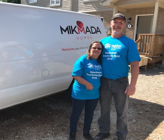 Two people in blue shirts stand next to a white Mikmada Homes van.