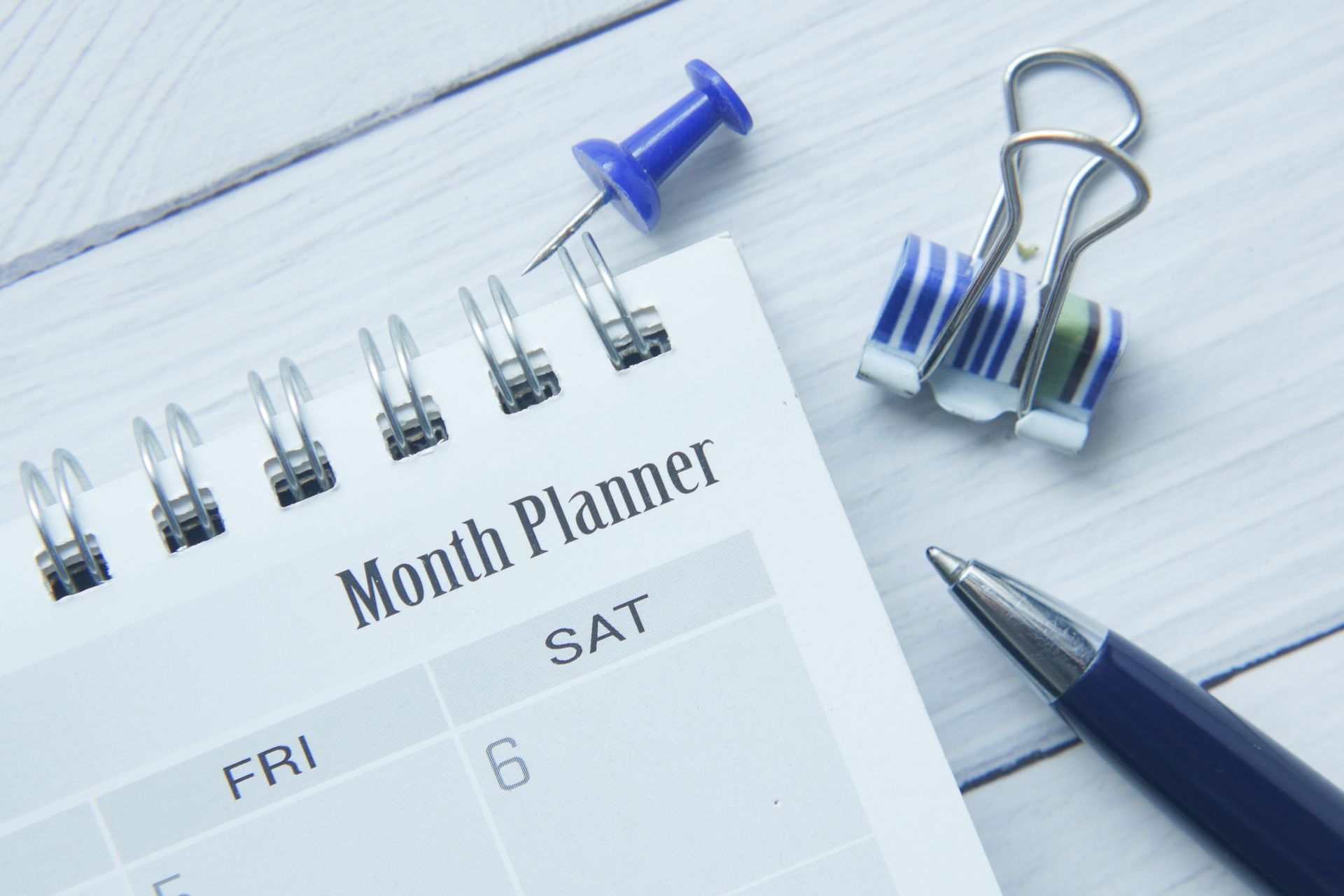 Month planner, push pin, binder clips, pen on light wood.