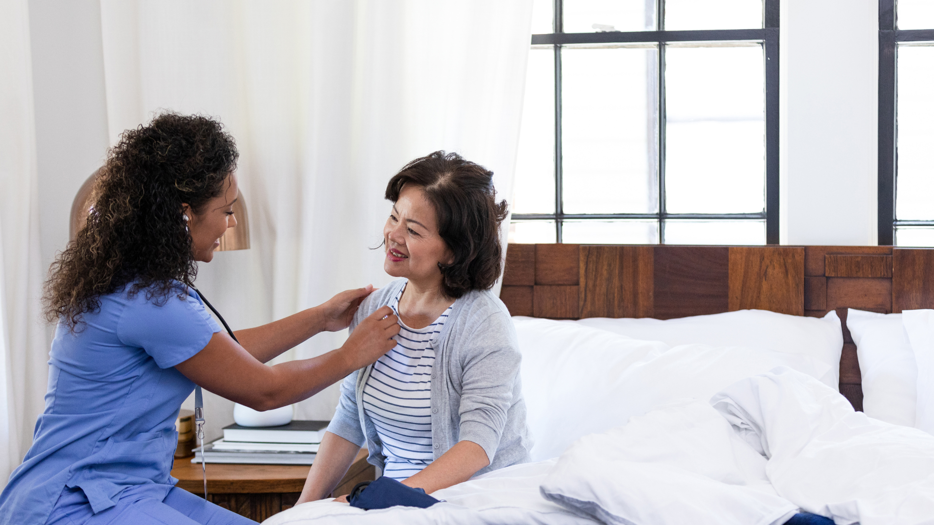 Caregiver listens to patient's chest with stethoscope in a bedroom.