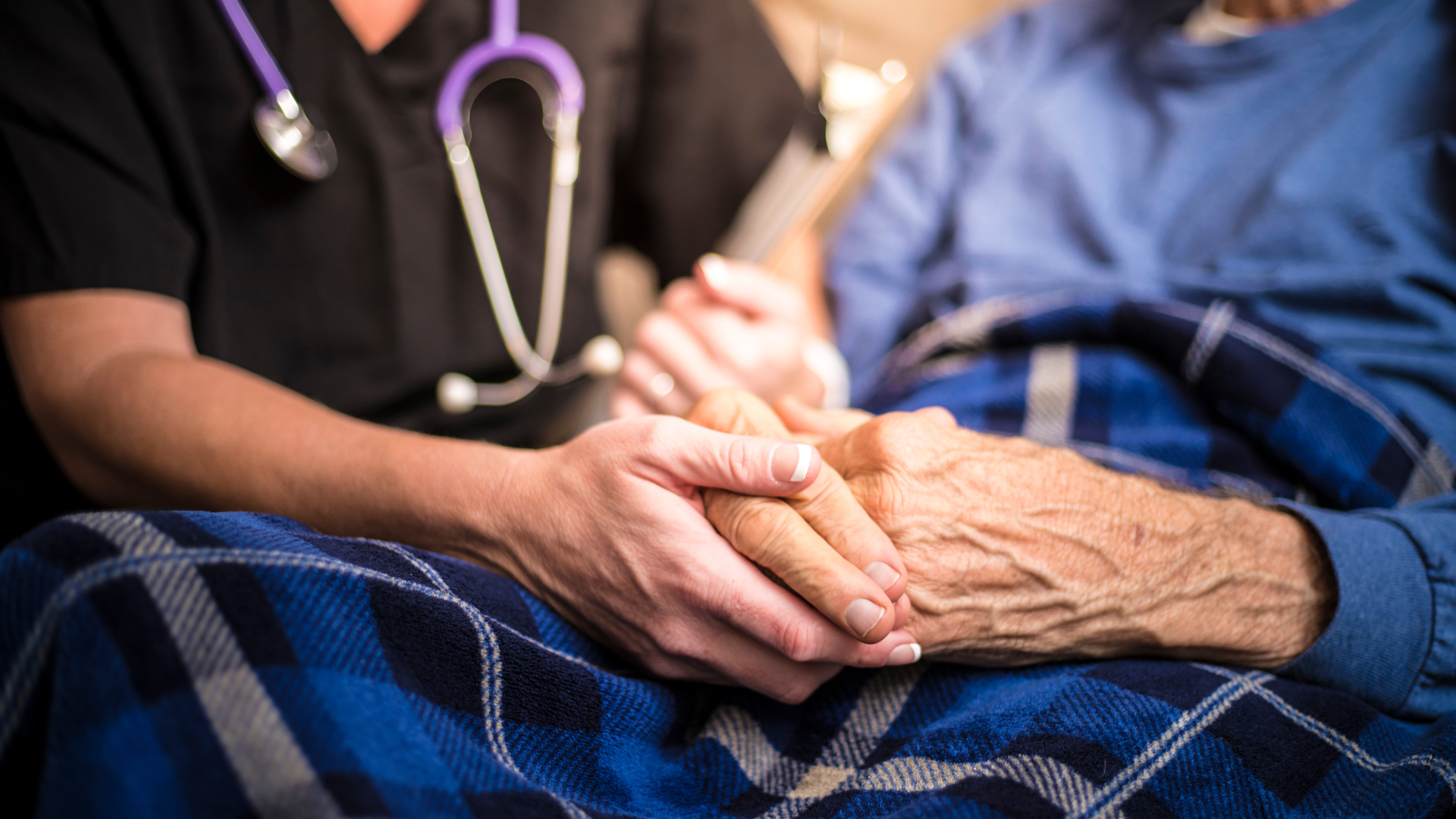 A caregiver in black scrubs and a stethoscope holds the hand of an elderly person, both resting on a blue plaid blanket.