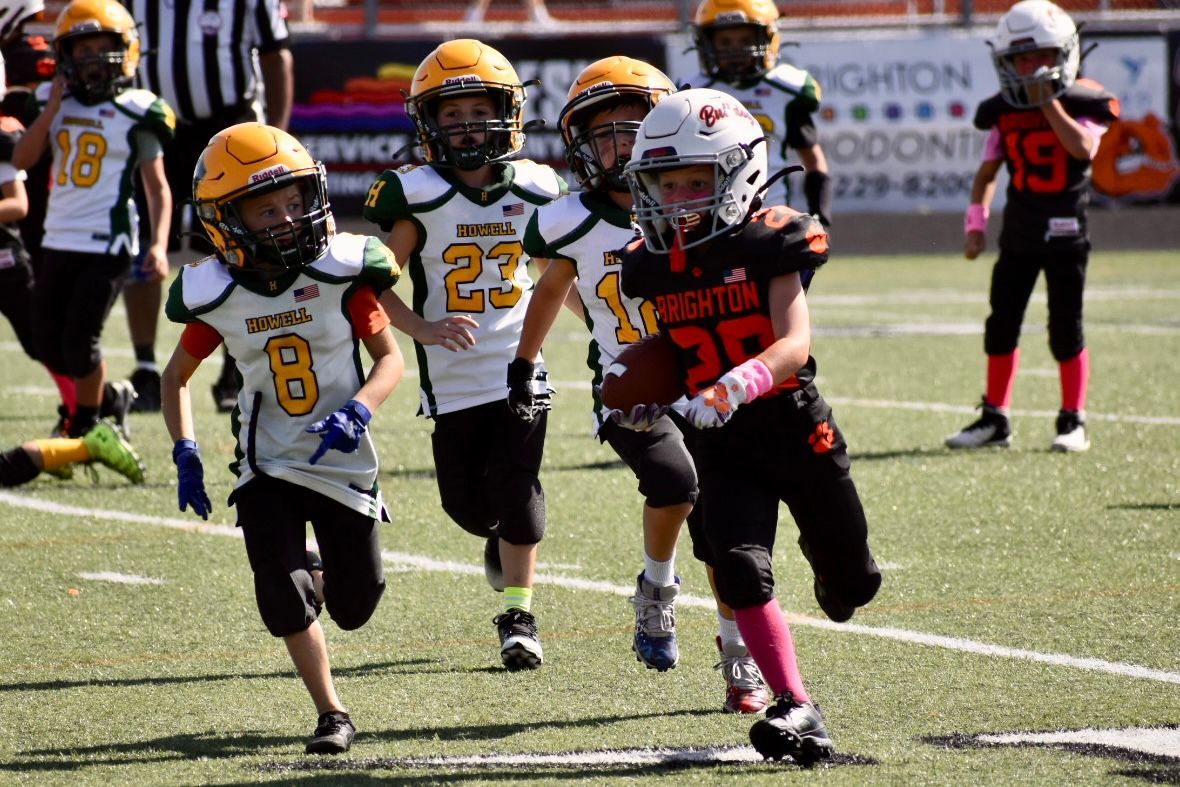 Youth football players sprint on a field during a game, with one in white and gold running ahead.