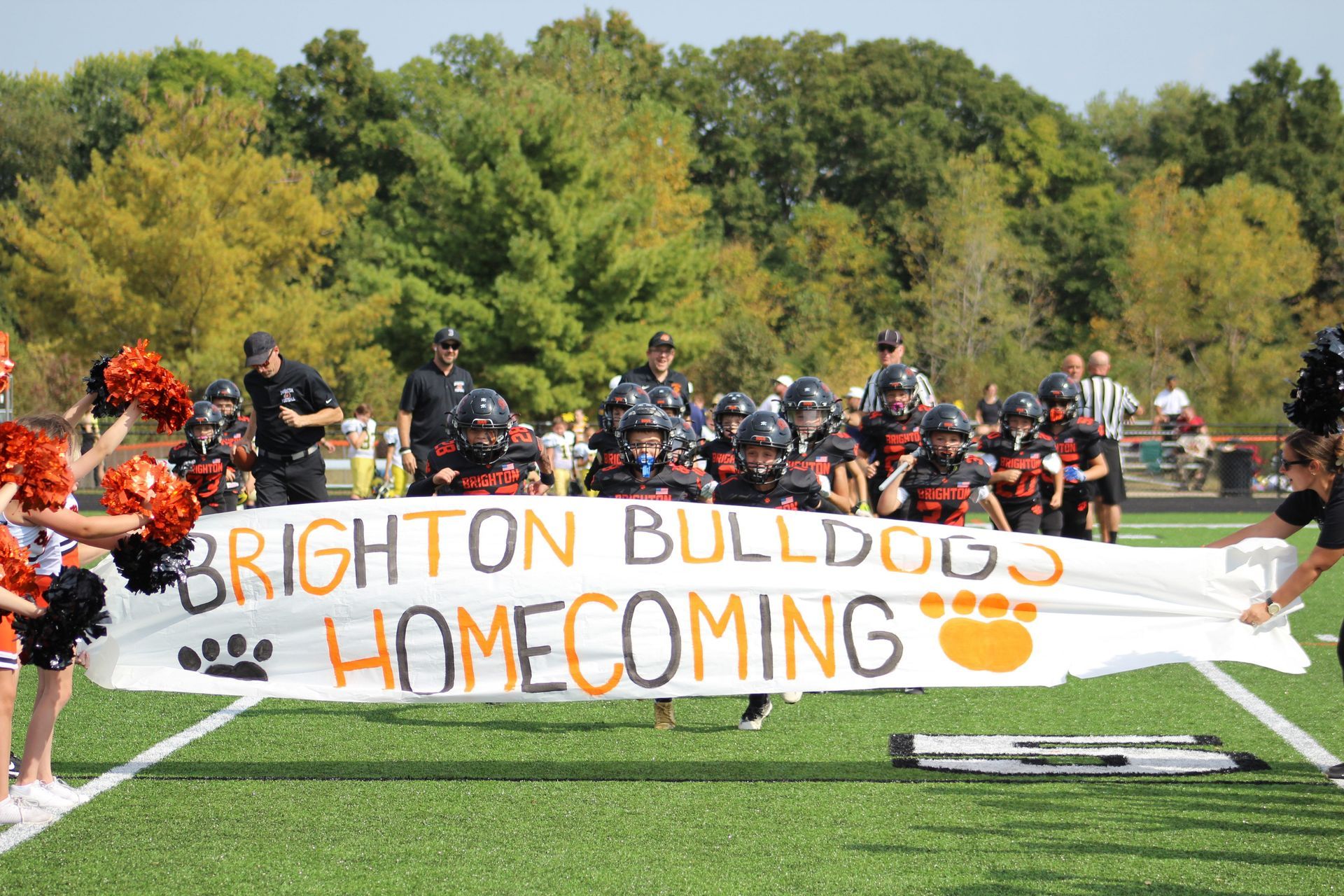 Football players huddle on a field near the goalpost, with coaches and trees in the background