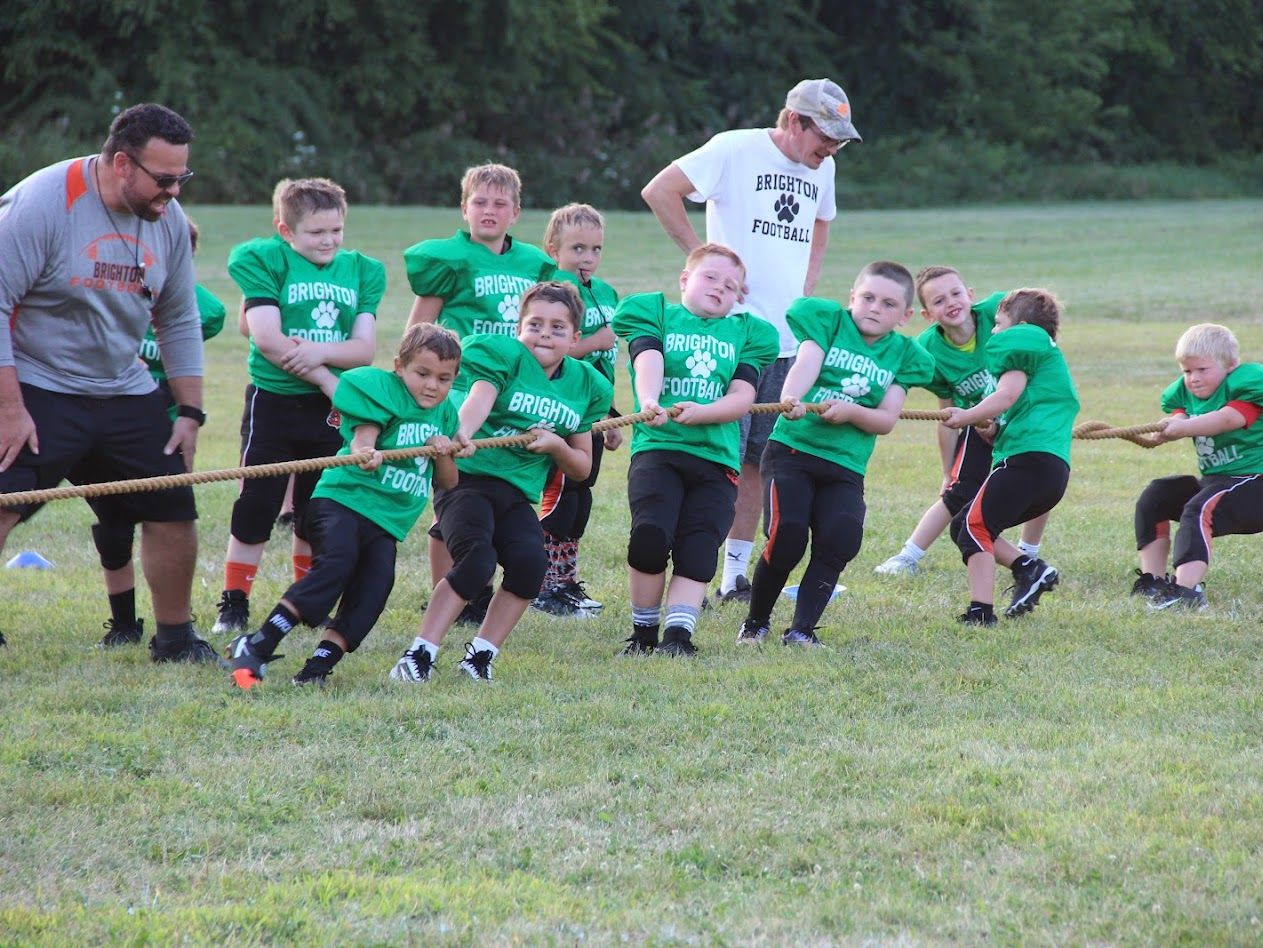 Children in green jerseys play tug-of-war on a grassy field with coaches nearby