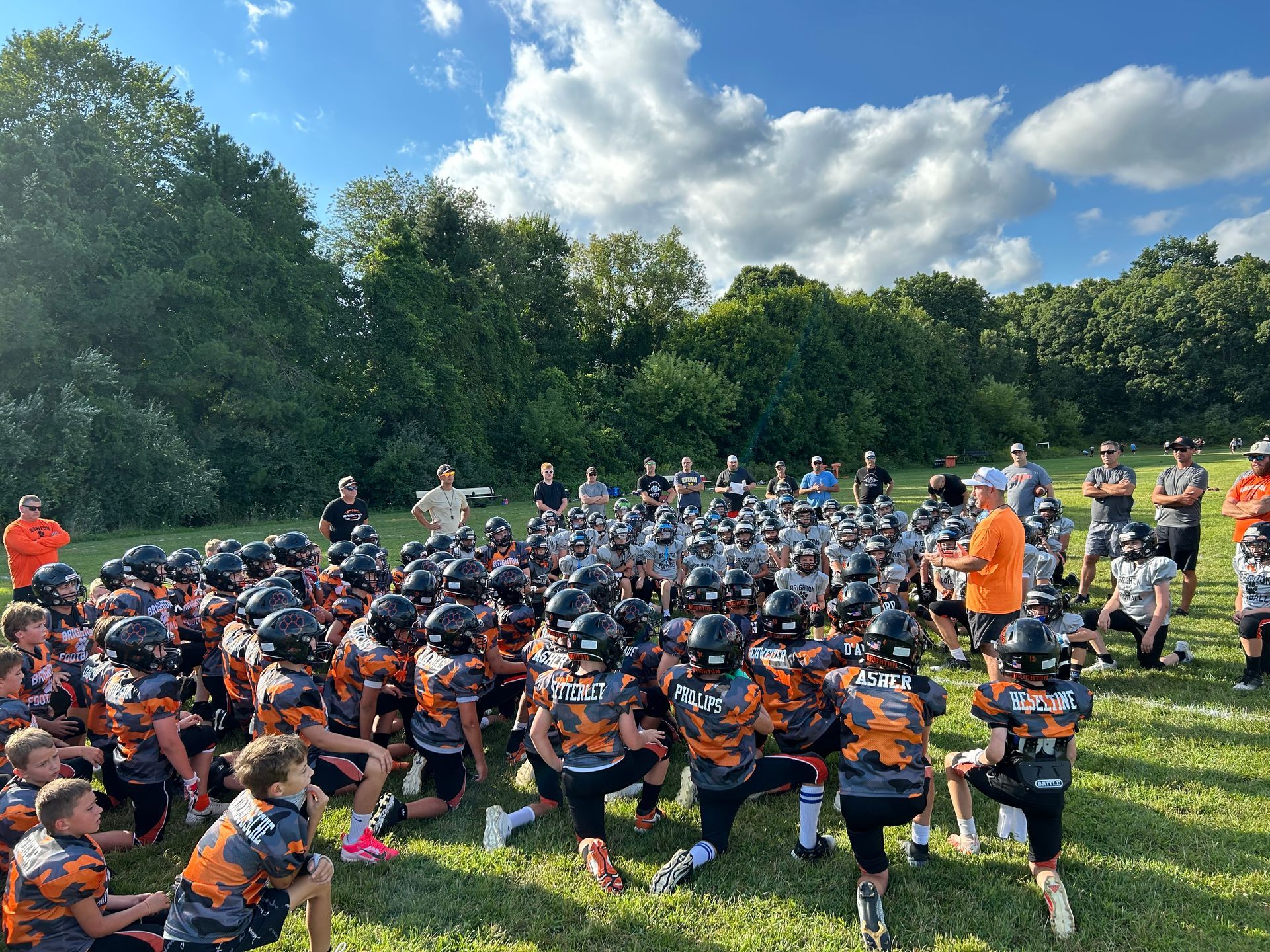 Youth football players in orange and black huddle on a field during practice.