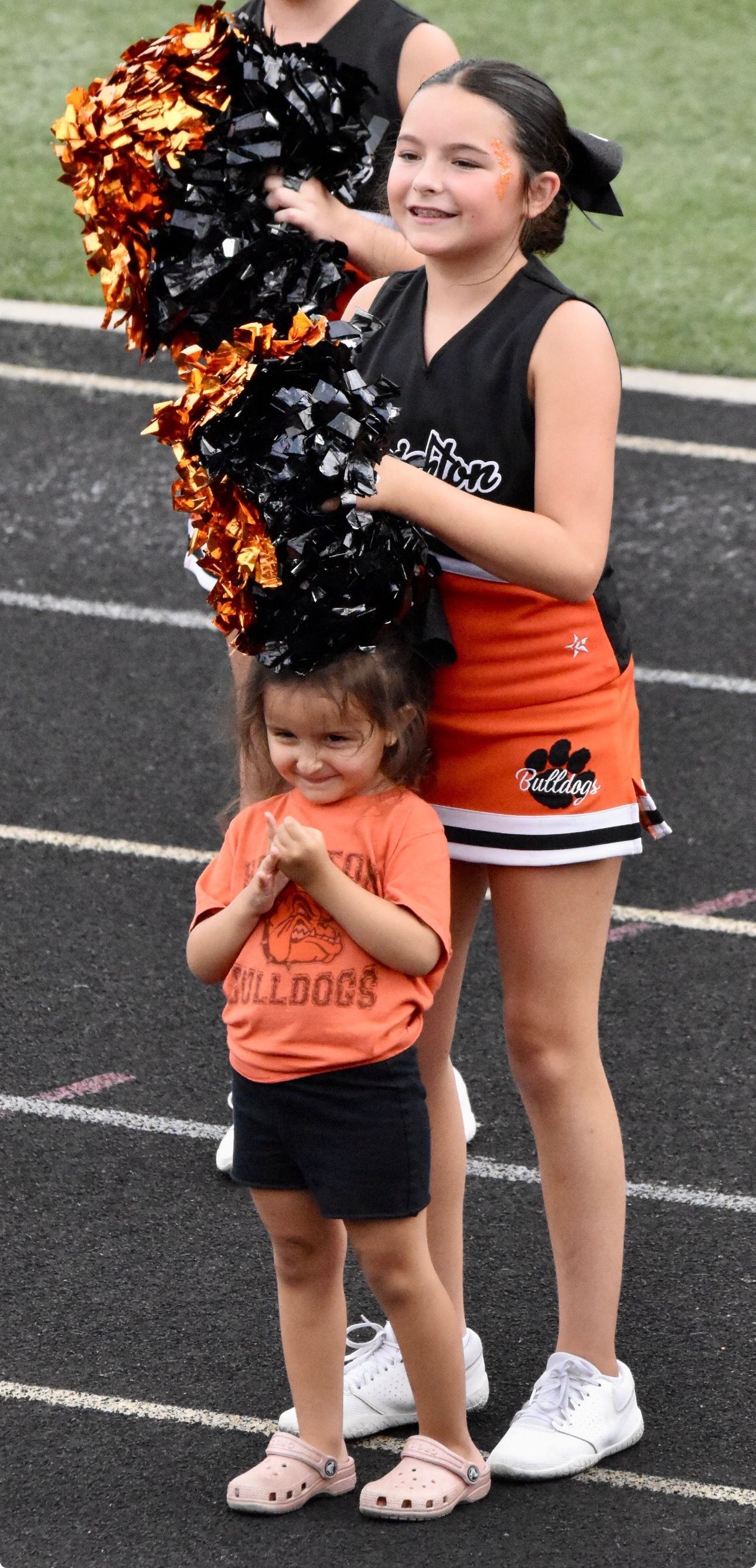 Youth football team posing on a field, players in black and orange uniforms with cheerleaders in front.
