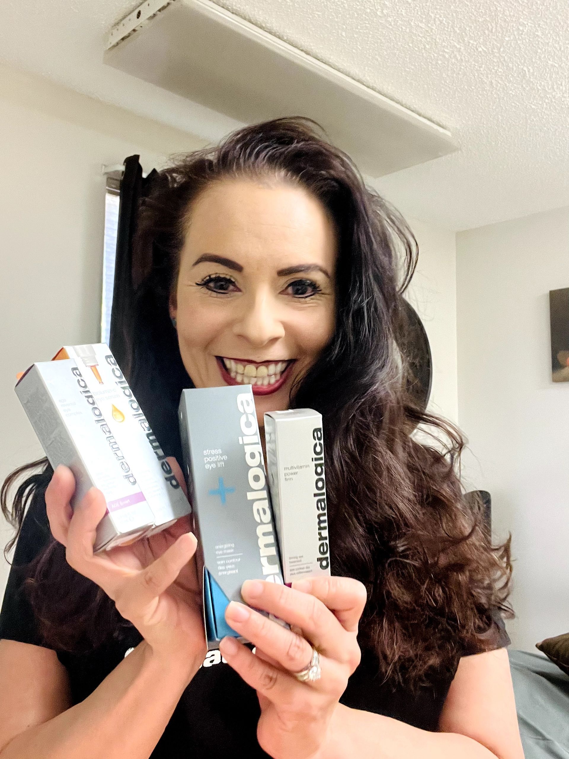 Woman with dark hair holding skincare boxes, smiling widely. Indoors.