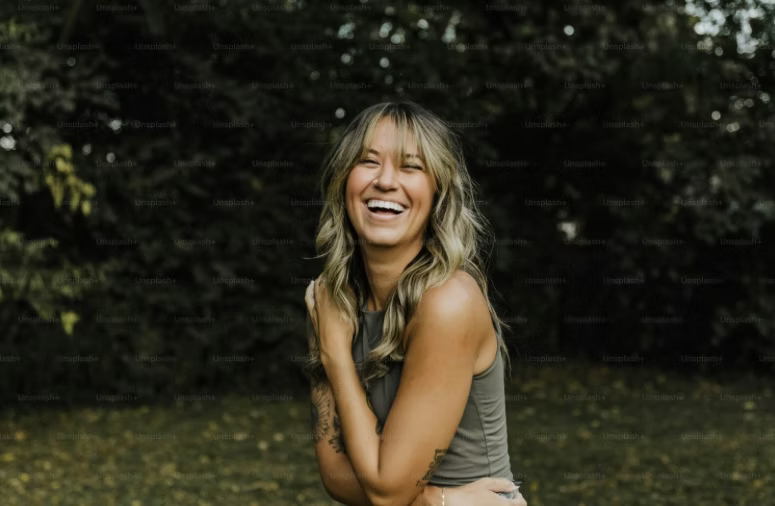 A woman is laughing in a park with her arms around her waist.