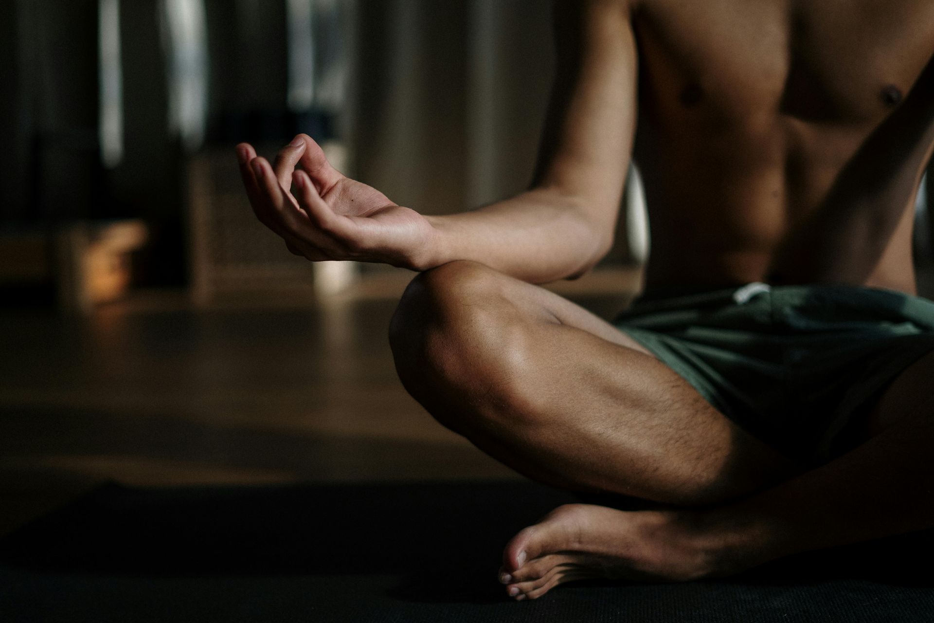Man meditating in lotus position, shirtless, indoors. Dark green shorts, hand gesture, focused.