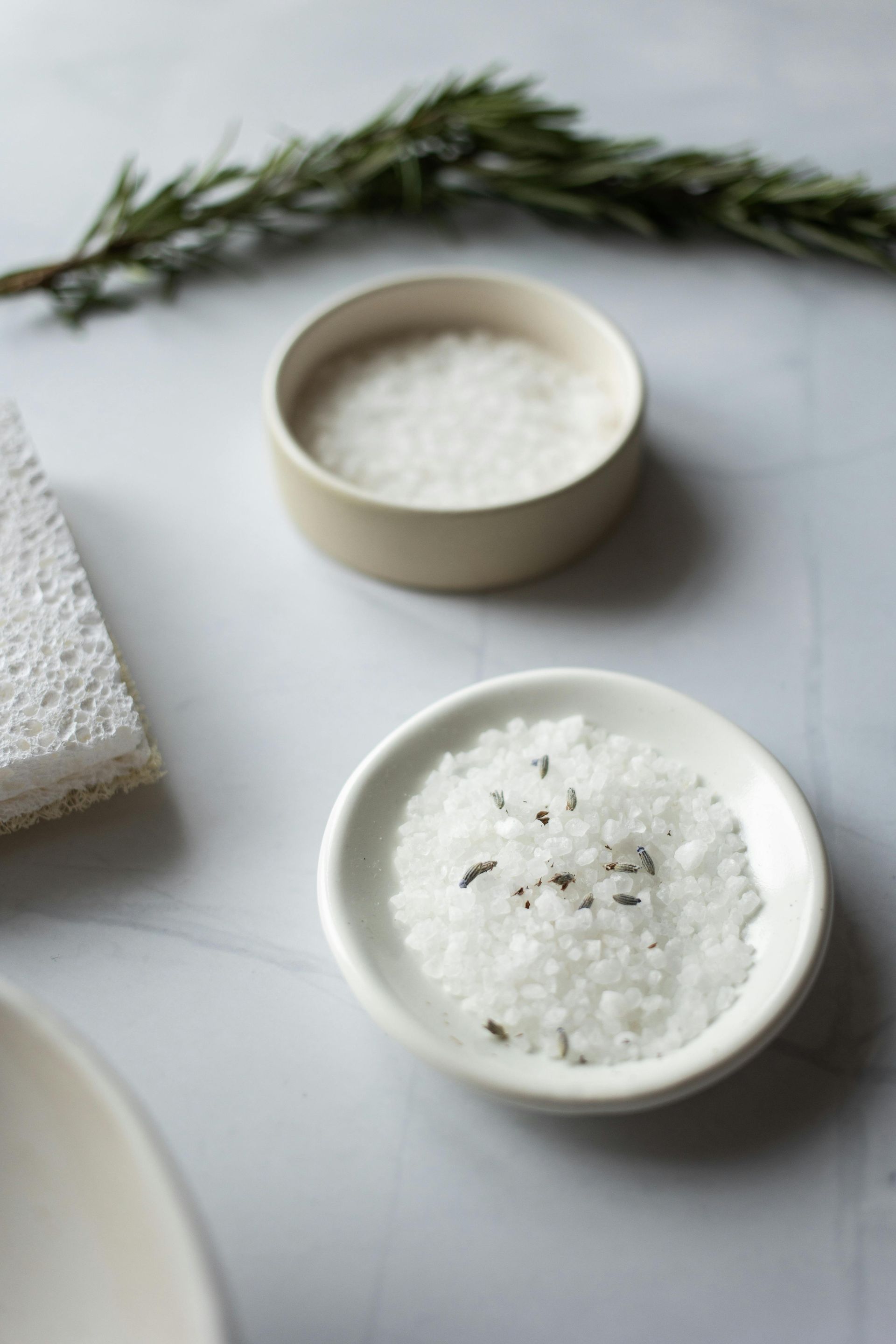 Two small bowls of salt, one with rosemary, on a white surface, with a sprig of rosemary and a sponge.